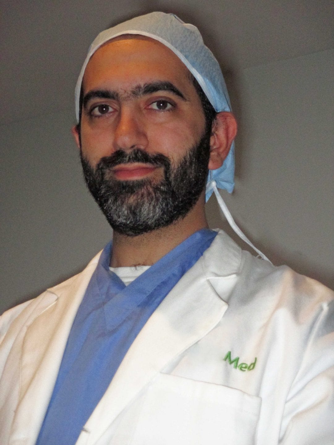 A man wearing a white lab coat, blue scrubs, and a surgical cap stands in front of a plain background.