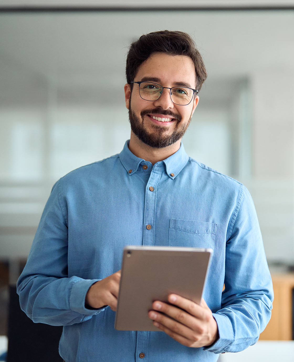 A man in a blue button-up shirt and glasses is standing indoors, holding a tablet and smiling at the camera.