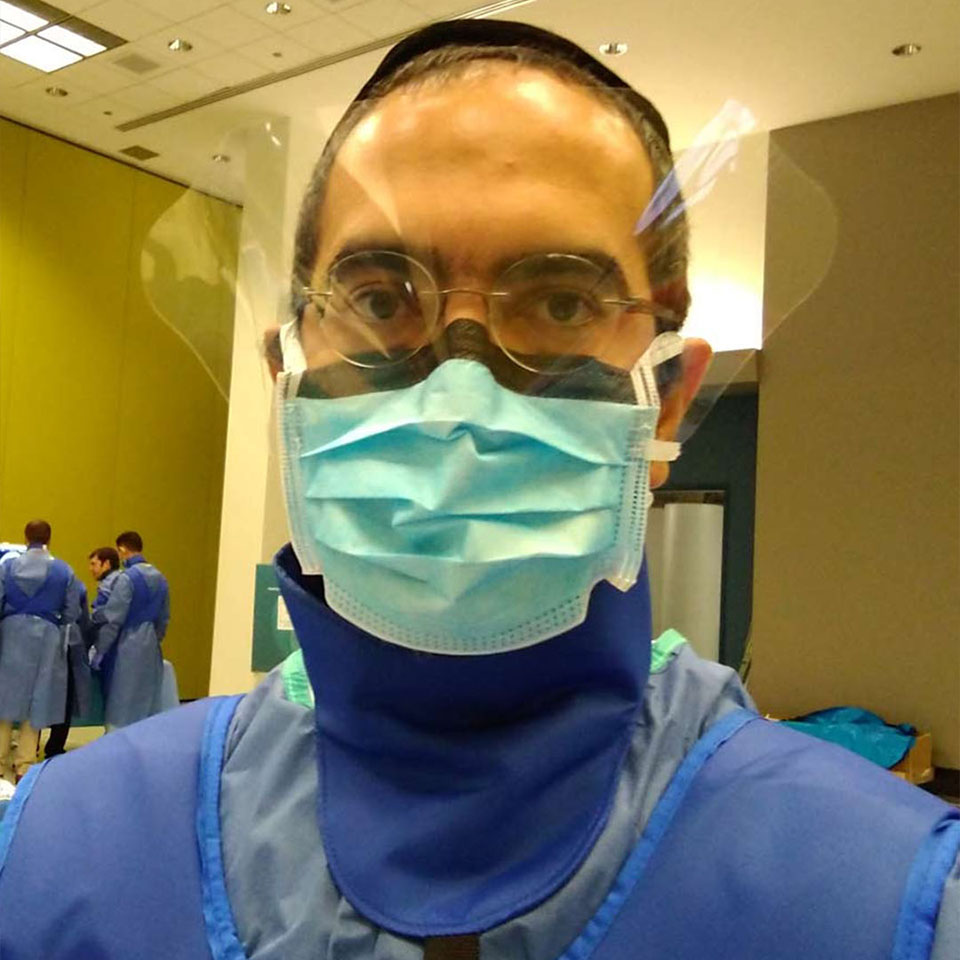 A person wearing protective gear, including a face shield, glasses, and a surgical mask, stands indoors. Other similarly dressed individuals are visible in the background.