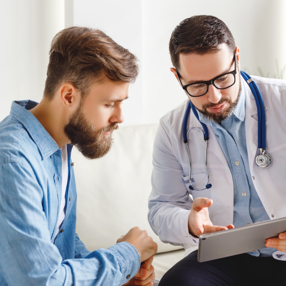 Medical practitioner in glasses showing test results to man and explaining diagnosis while sitting on couch in office of modern clinic