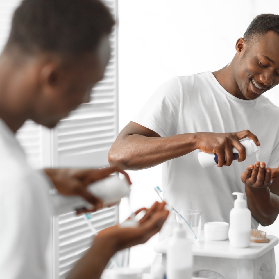 Cheerful African American Man Using Shaving Foam Standing Near Mirror In Bathroom At Home. Male Morning, Facial Skincare Cosmetics And Self-Care Routine For Men Concept. Selective Focus