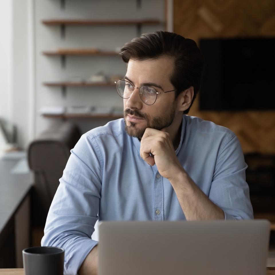 Thoughtful businessman in glasses looking at window from workplace with laptop, thinking over challenges, work issues, problem solving, making decision, feeling doubts, pondering on future vision