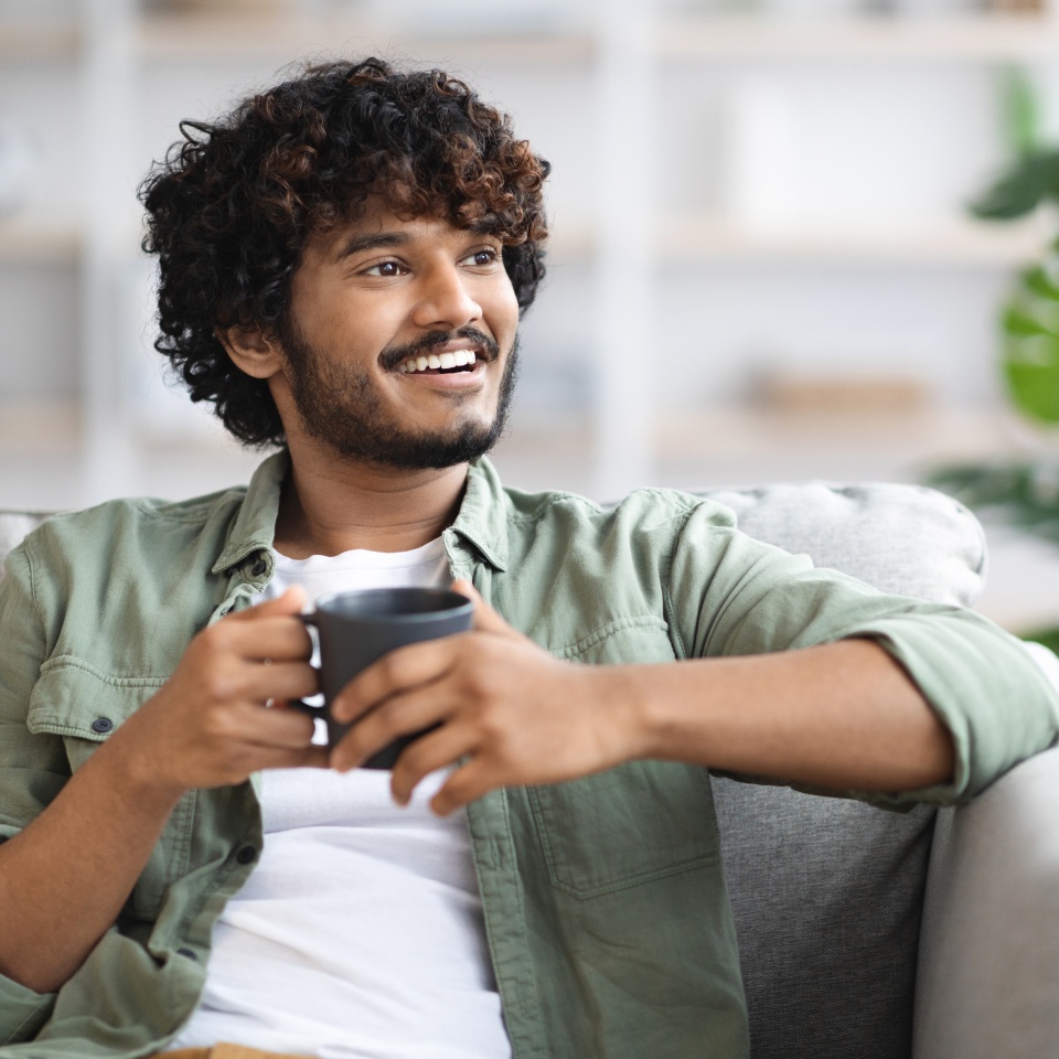 Handsome young guy with curly hair sitting on couch in cozy living room, holding tea mug, looking at copy space and smiling, enjoying morning coffee at home. Domestic lifestyle concept