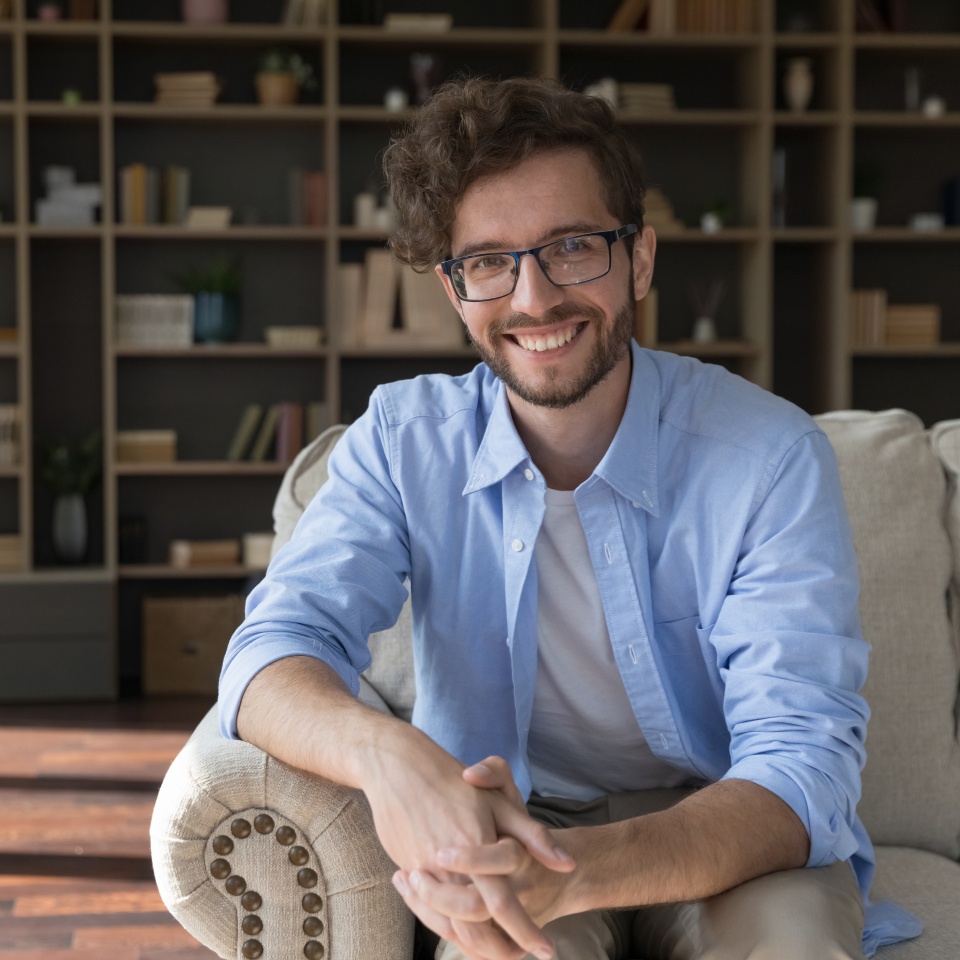 Happy handsome young 20s business man in glasses sitting on couch at home with bookshelves in background, looking at camera, smiling. Happy blogger, freelancer in eyeglasses head shot portrait