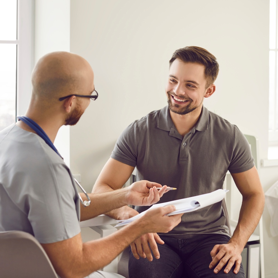 Portrait of a happy smiling young man patient in medical office listening to doctor holding report file with appointment and giving consultation during medical examination in clinic.
