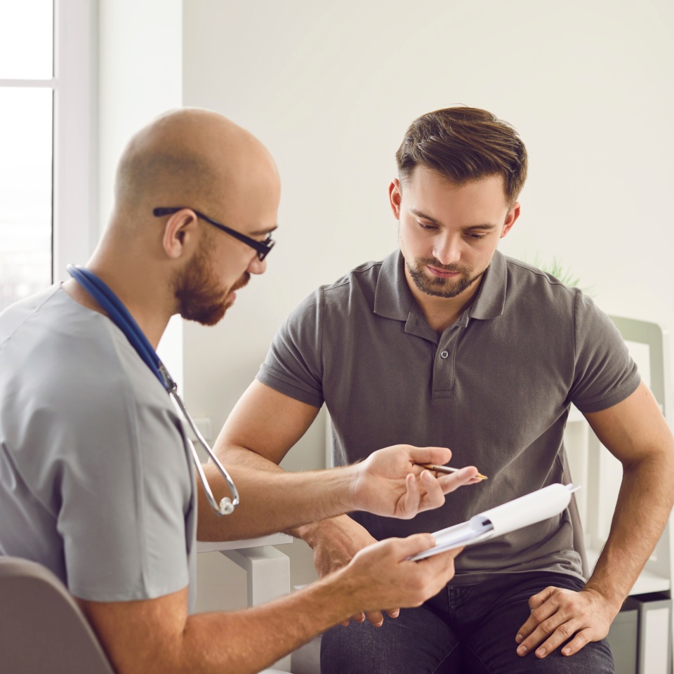 Doctor talks to his patient after a medical examination at the clinic. Young male physician shows a clipboard with the results of some medical tests they did and explains what this all means