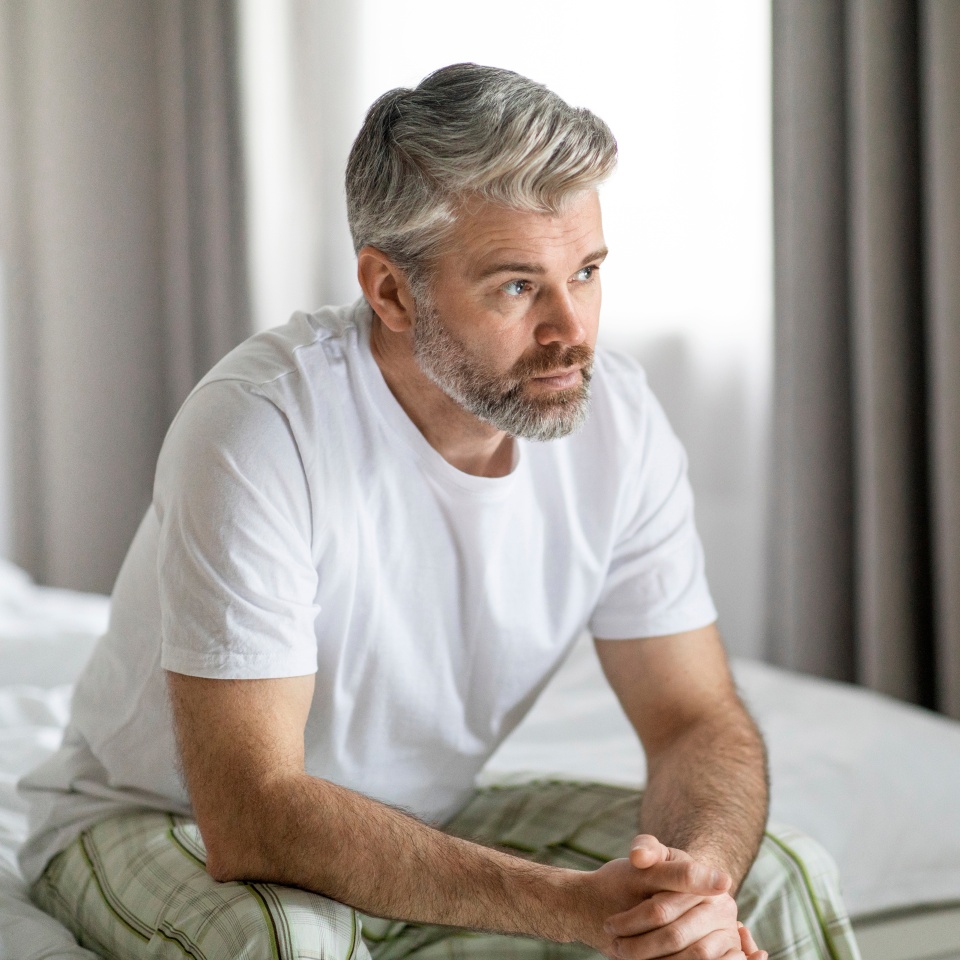 Pensive thoughtful unhappy handsome grey-haired bearded mature caucasian man wearing pajamas sitting on bed at home, looking at copy space, feeling lonely, going through divorce