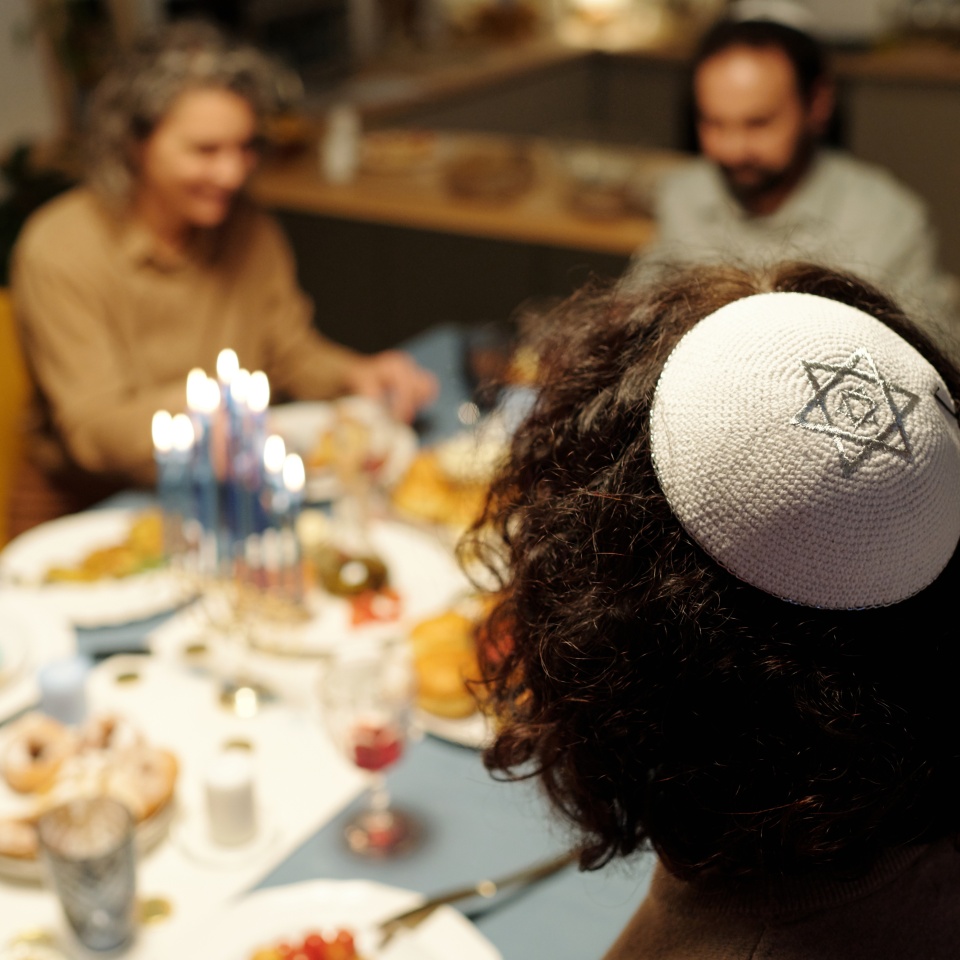 White yarmulke with silver star of David on head of young Jewish man sitting by served table with homemade food and drinks