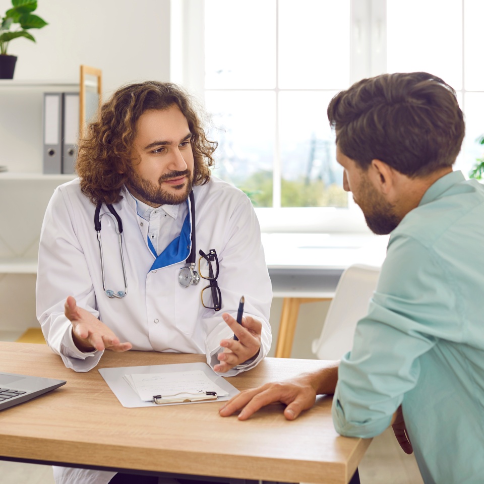 Doctor and patient having conversation. Male physician in white medical lab coat sitting at desk with laptop, talking to worried man, giving advice, explaining importance of changing his lifestyle