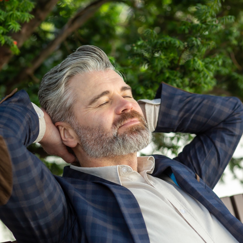 Senior professional man in business attire takes a peaceful break outdoors, reflecting with eyes closed amidst greenery.