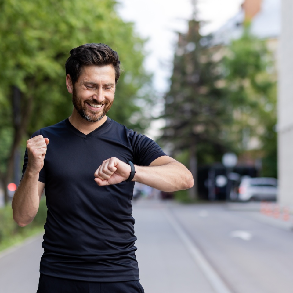 Happy young man doing sports and running, standing on city street, looking at wrist tracker and showing success gesture with hand.