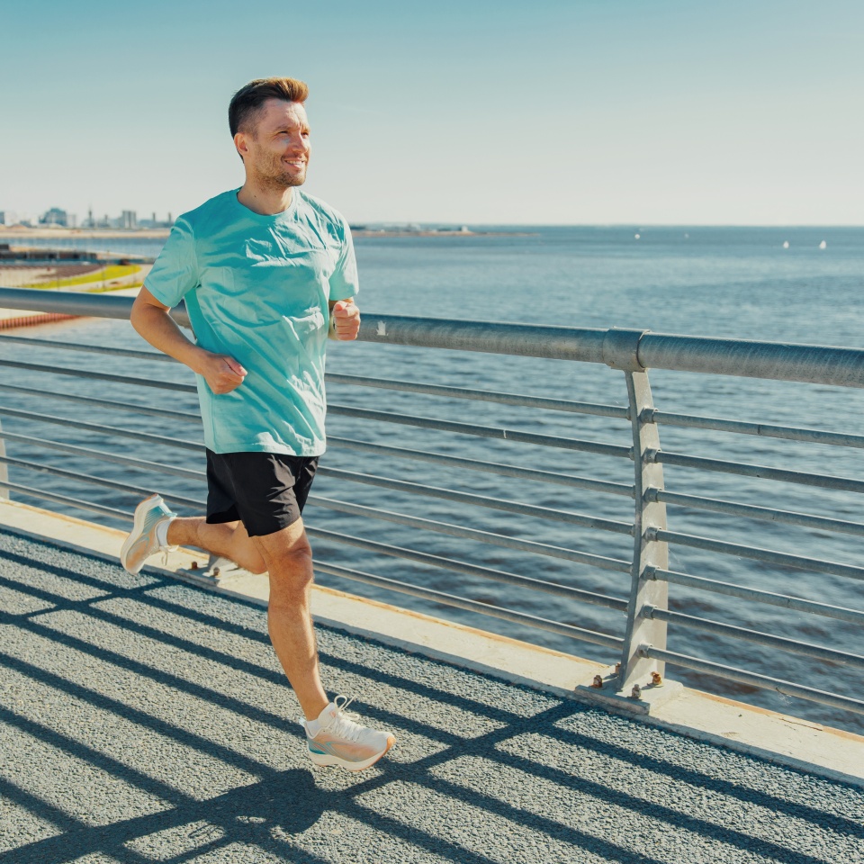 A man in a turquoise shirt jogs on a waterfront pathway, enjoying a clear, sunny day with a serene ocean view.