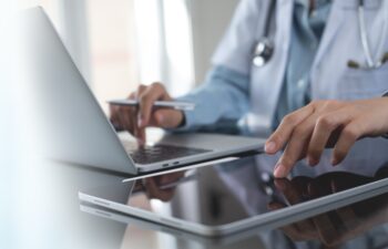 Doctor using digital tablet and working on laptop computer at doctor's office in hospital, close up. Healthcare and medicine, medical technology background