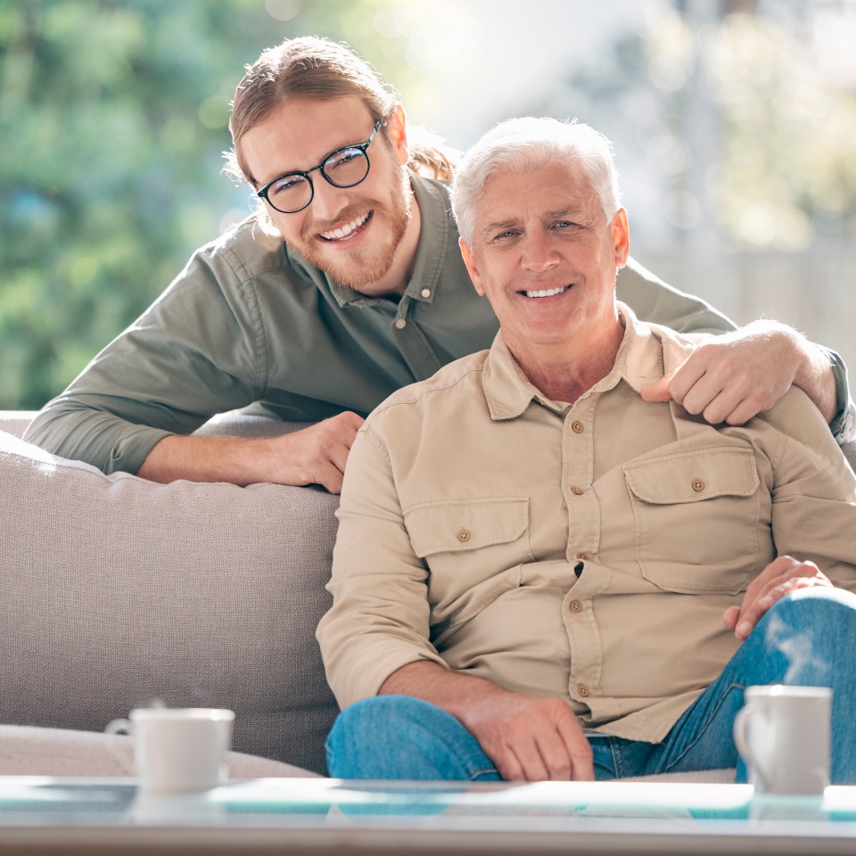 Happy, portrait and man with senior dad on sofa to relax together for fathers day, care or support. Smile, family and proud elderly male parent with person or son for bonding in living room in home