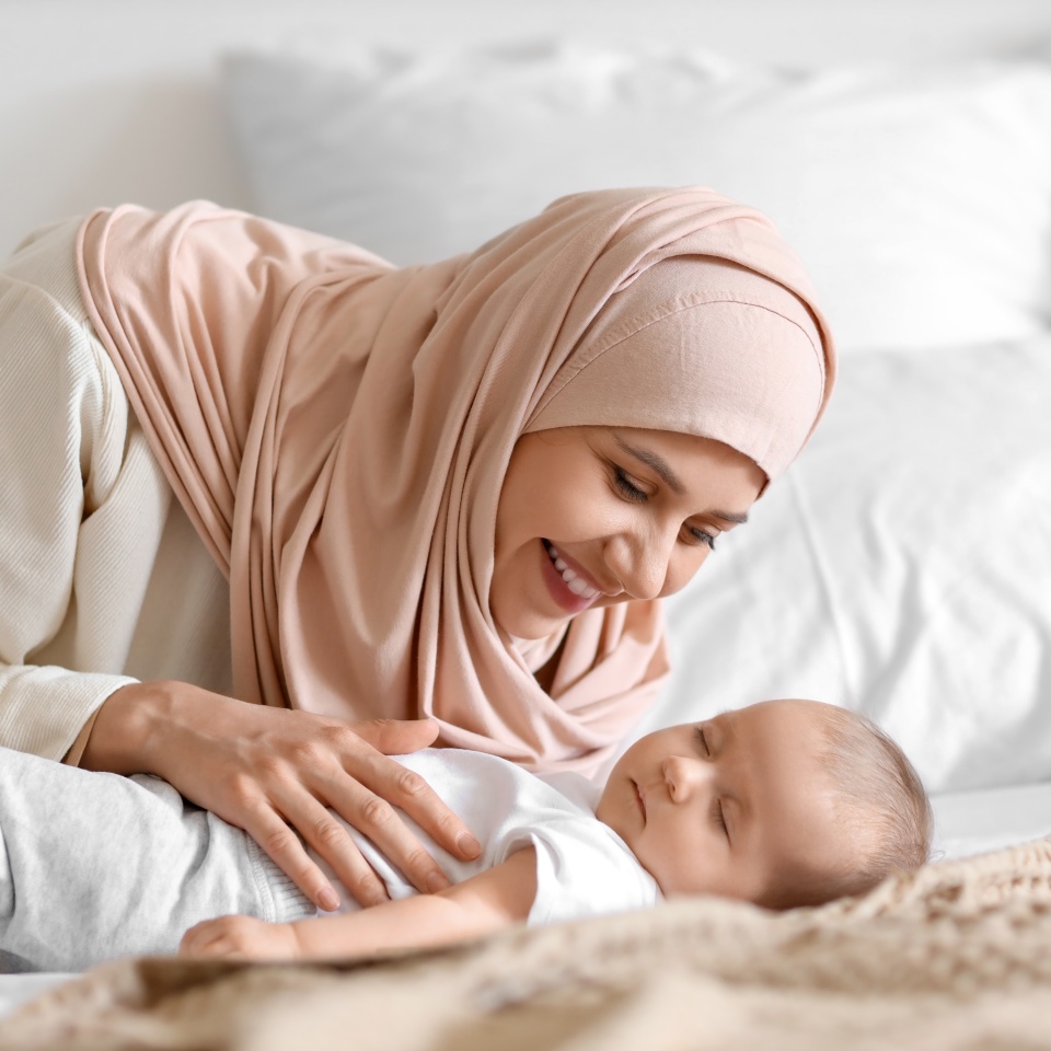 Happy Muslim mother in hijab and cute little baby sleeping on bed at home