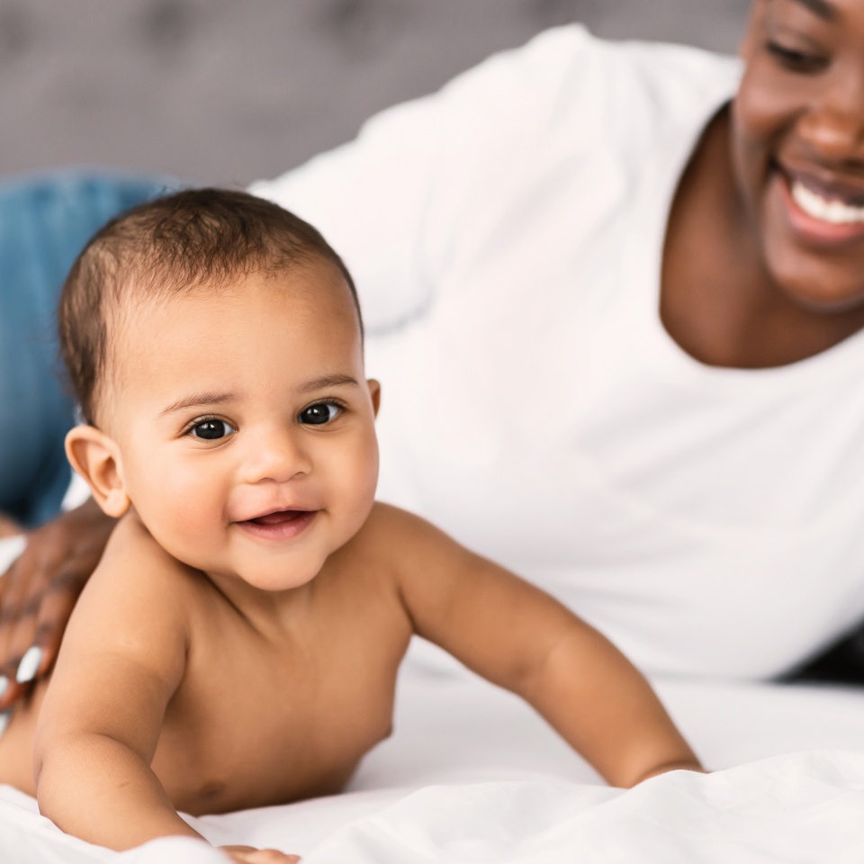 Happy Loving Family. Portrait of smiling African American mom taking care of her adorable small black baby who lying on tummy on the white blanket at home. Infant kid crawling on bed in bedroom