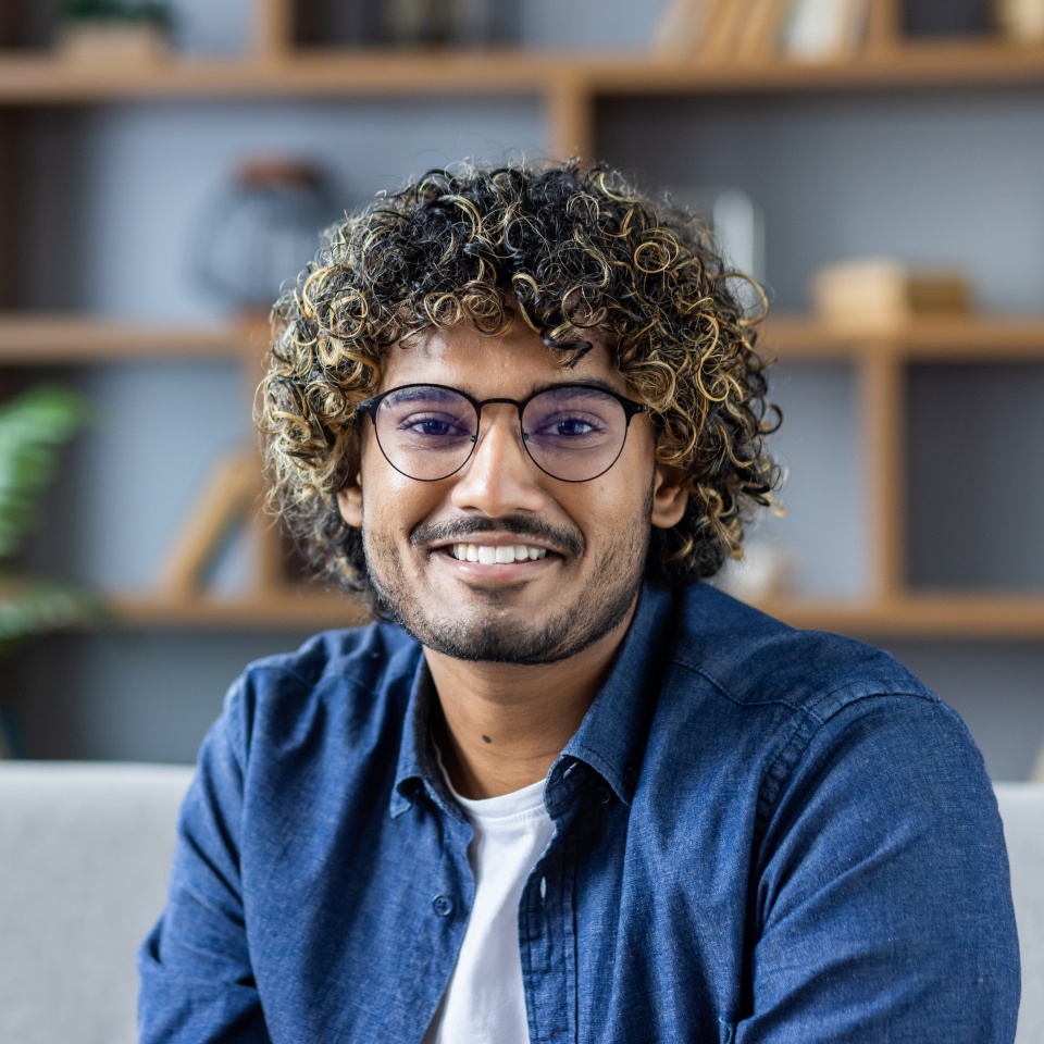 A young man with curly hair and glasses is sitting on a sofa, smiling warmly. He is wearing a casual blue shirt, creating a friendly and approachable vibe in a modern living area.