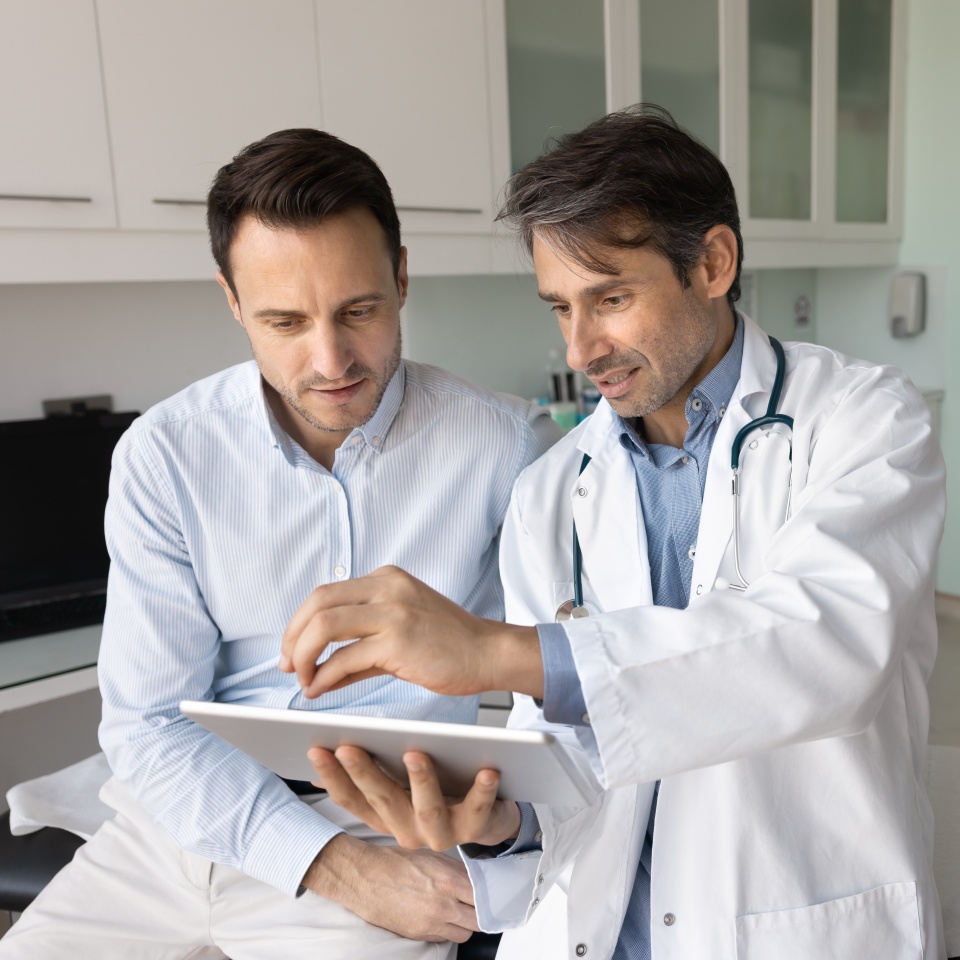 Focused middle aged Latin doctor man showing examination reports to patient, explaining ultrasound scanning test result, pointing at tablet screen, using electronic technology for work with patient