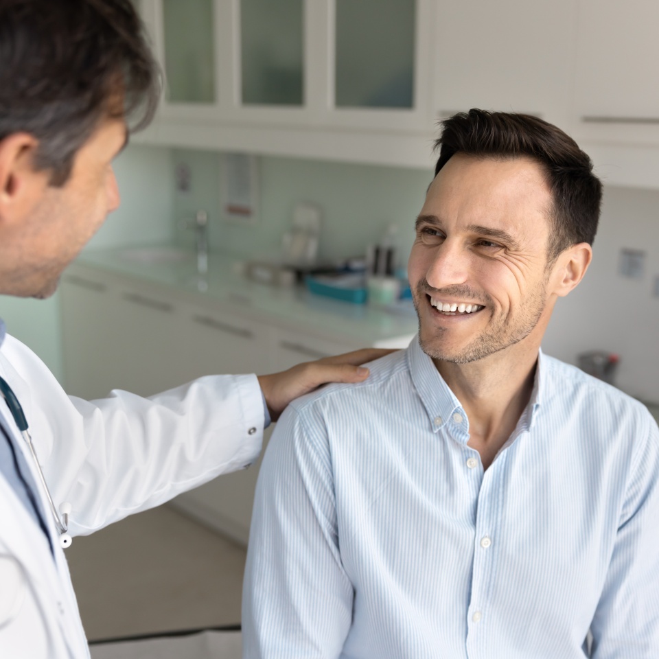 Cheerful patient man getting supportive shoulder touch gesture from doctor, sitting on couch in examination room, visiting medical specialist for checkup, learning good news about test results