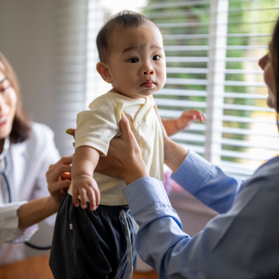 A woman is holding a baby while a doctor examines the baby. The baby is wearing a white shirt