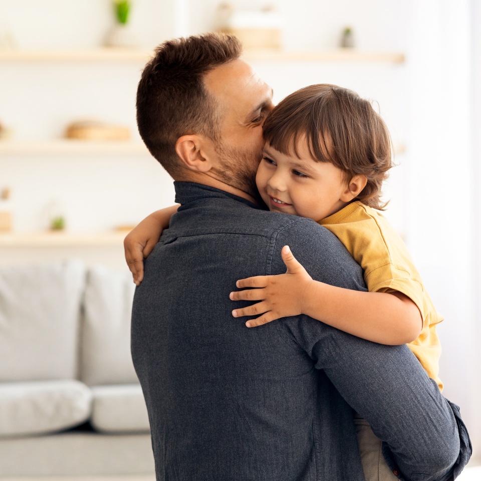 Cute little boy enjoying time with loving daddy, caring man embracing and kissing his lovely son, holding him on hands at home interior