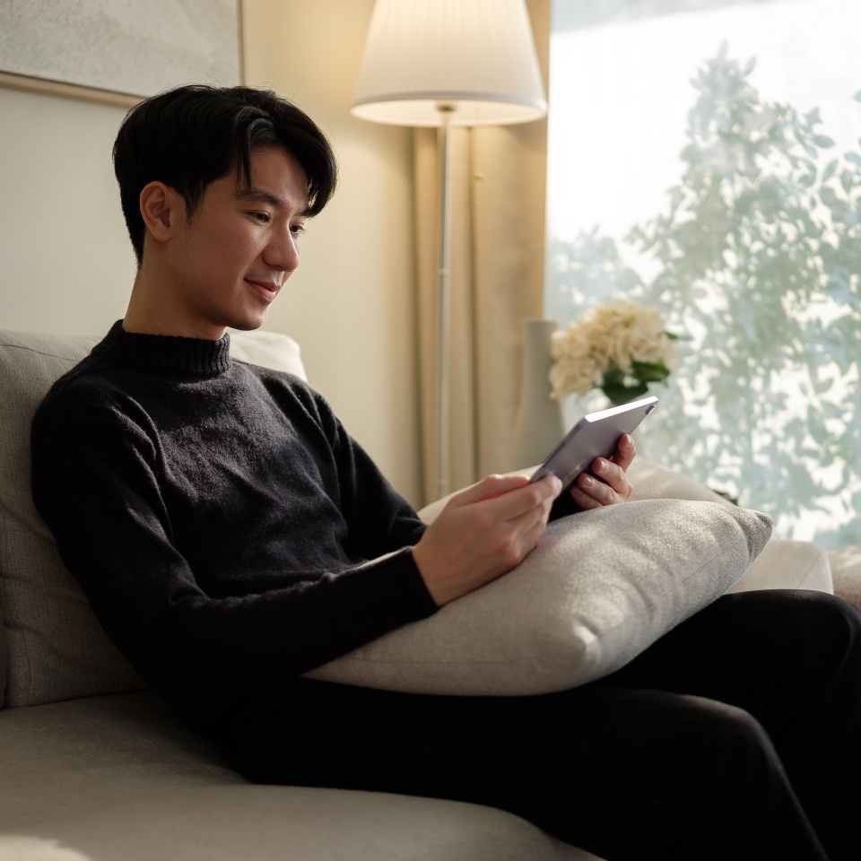 Young man in a cozy black sweater sits comfortably on a soft couch, holding a tablet..