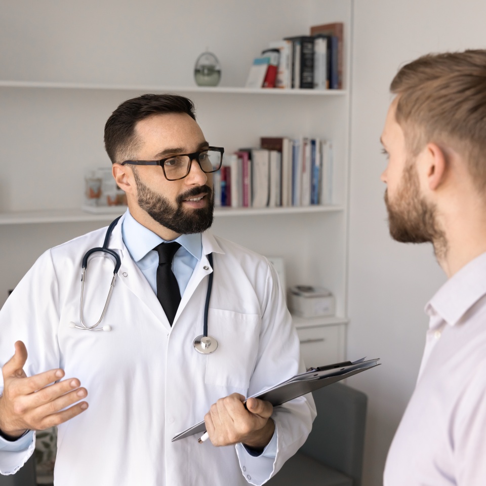 Confident male physician doctor providing medical advice to patient, talking to young man on consultation meeting in hospital examination room, explaining diagnosis, treatment