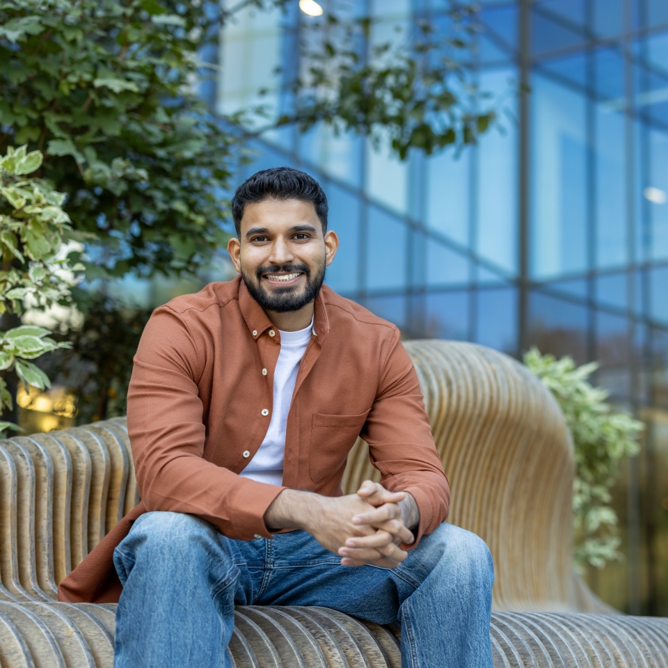 A smiling man of Indian descent sits on a modern wooden bench outdoors with a modern building in the background.