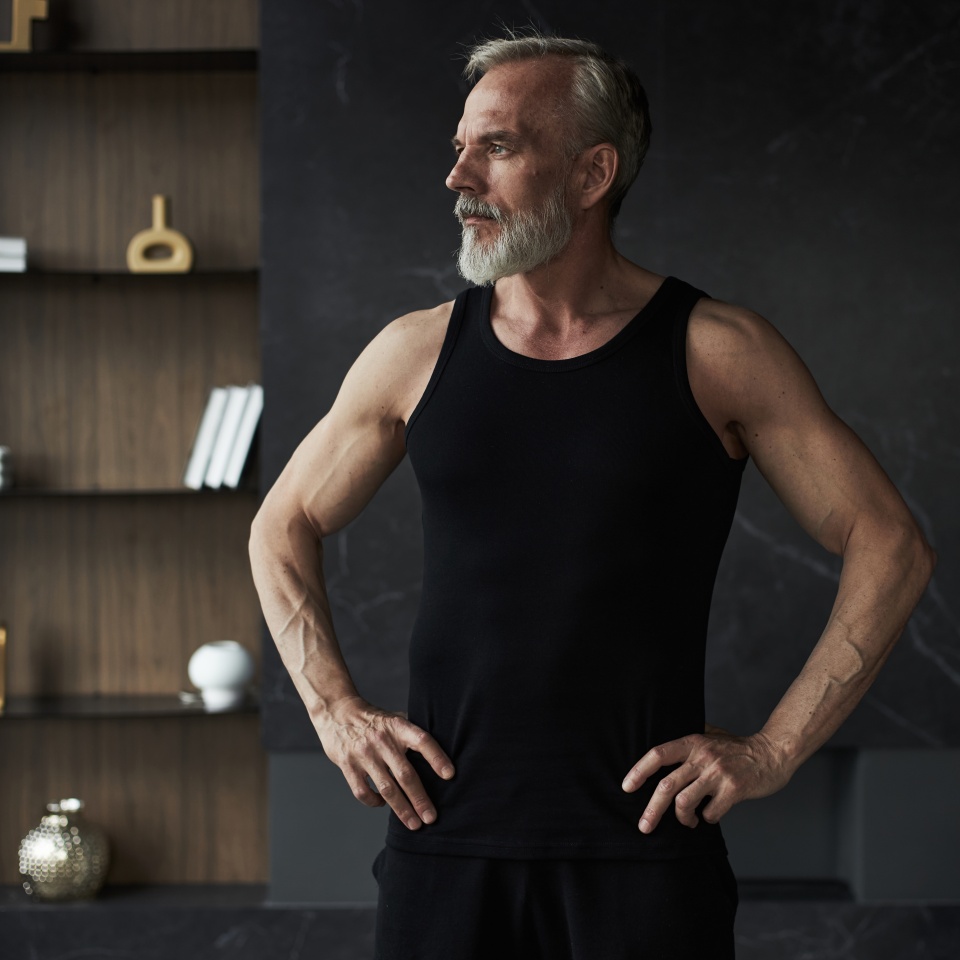 Middle aged Caucasian man standing with hands on hips looking away from camera showing muscular arms and confident posture in modern interior