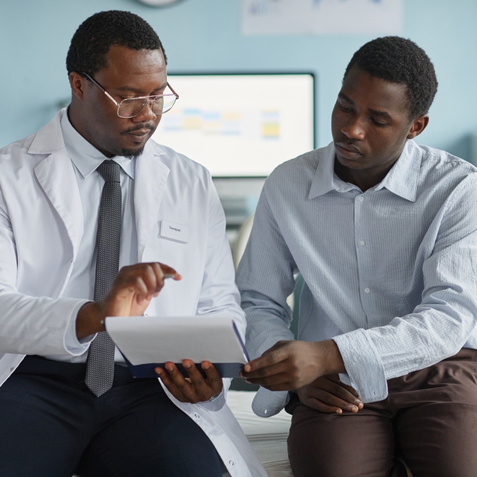 Black middle aged male doctor explaining medical information on digital tablet to Black young adult man patient during consultation in modern medical office