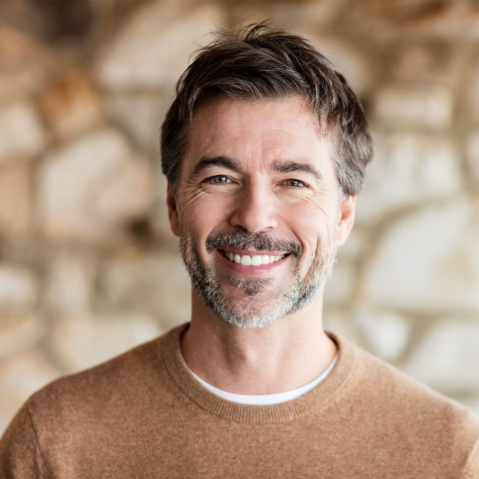 A man with short brown hair, a beard, and a mustache, wearing a tan sweater, smiles at the camera with a stone wall background.