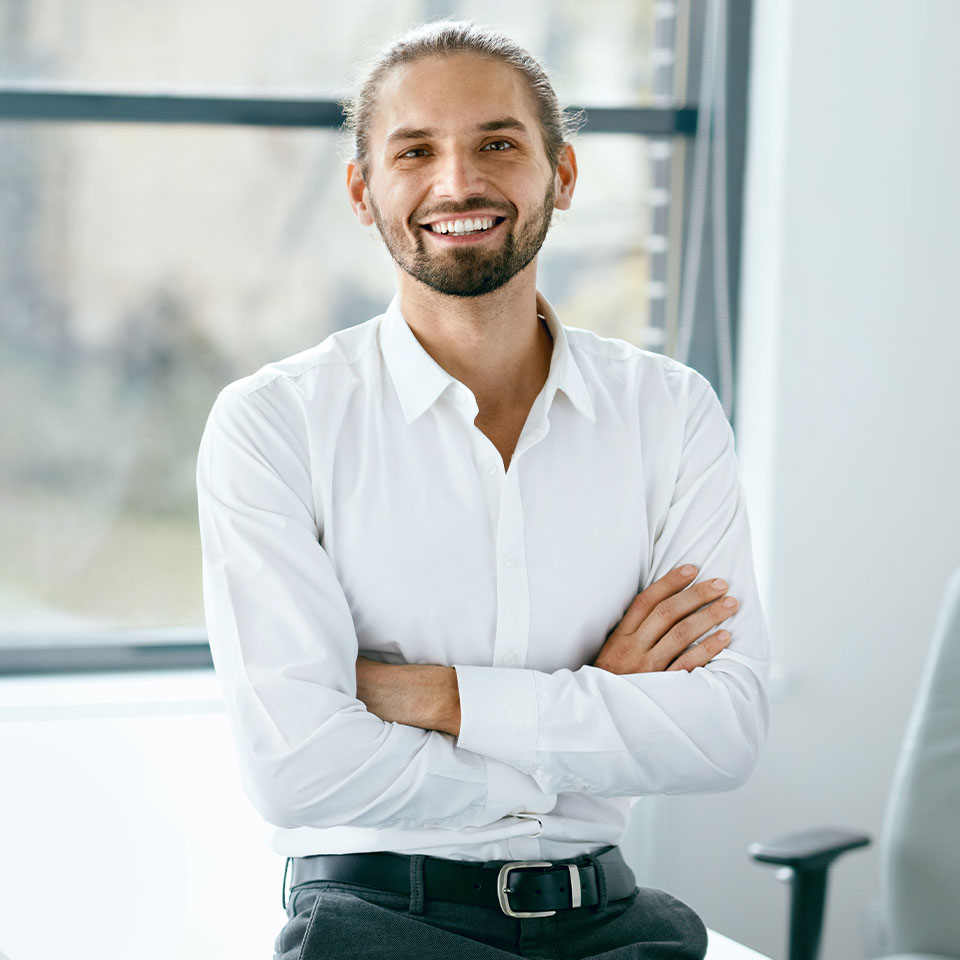 A man with long hair tied back, a beard, and mustache, wearing a white shirt, stands with arms crossed and smiles in a bright office setting.