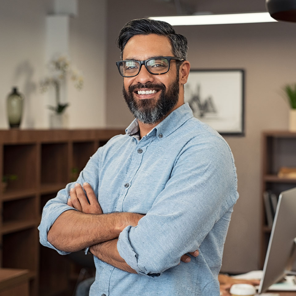 A man with glasses and a beard, wearing a light blue shirt, stands smiling with arms crossed in an office setting.