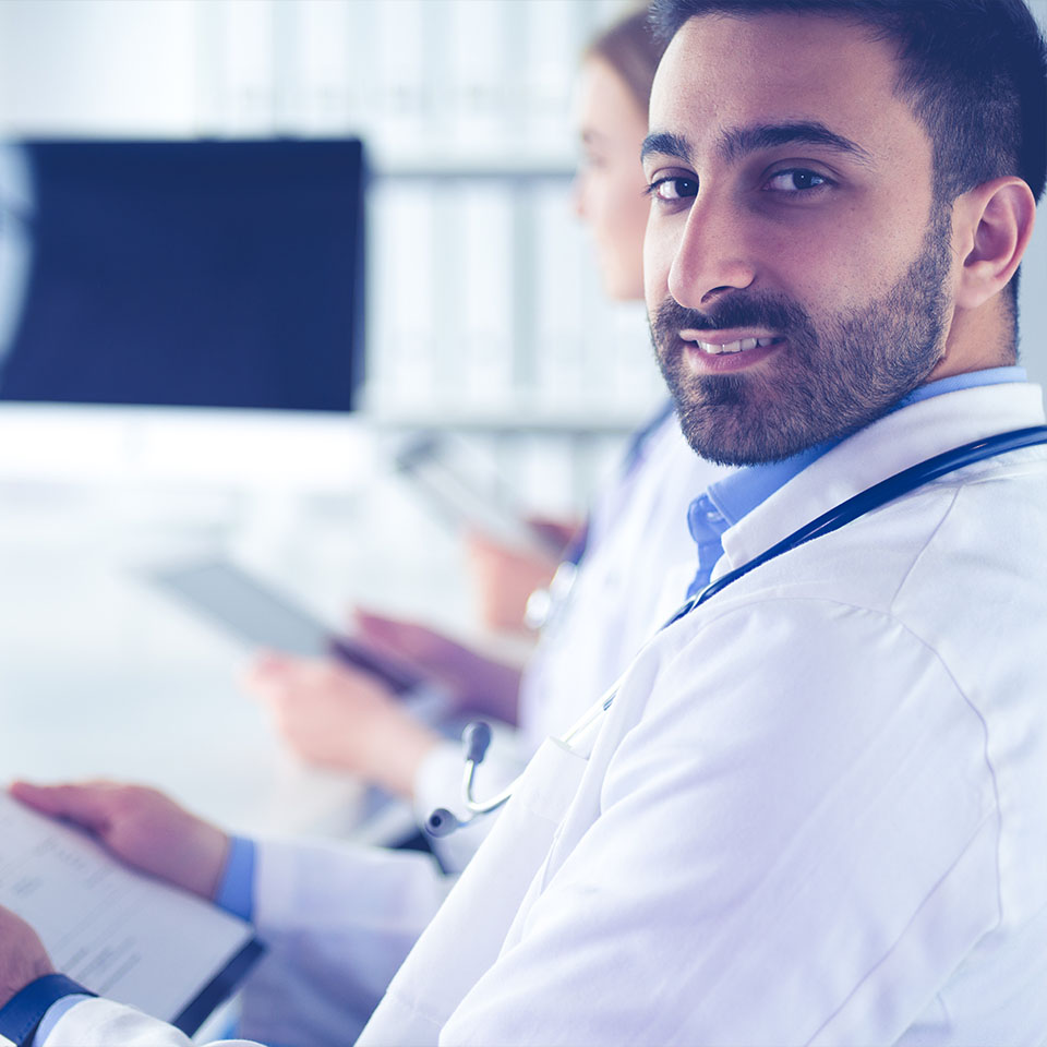 A male doctor wearing a white coat and stethoscope looks at the camera while holding a clipboard, with another medical professional working in the background.