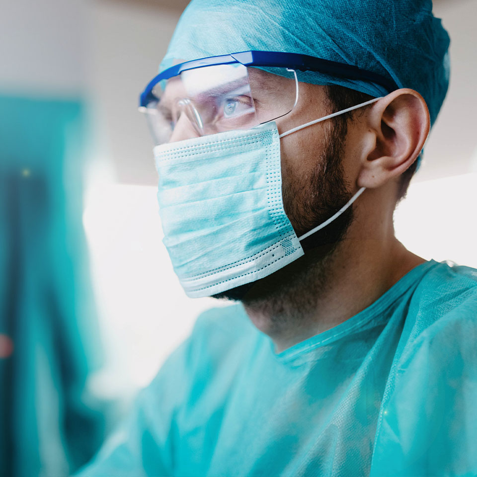 A healthcare worker wearing a surgical mask, protective glasses, hair cover, and a blue gown looks ahead in a clinical setting.