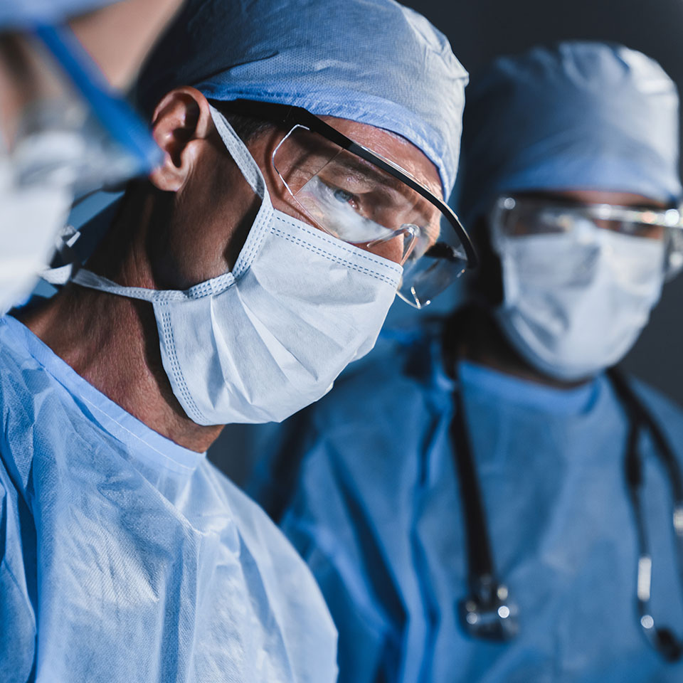 Several surgeons wearing surgical masks, scrubs, caps, and protective glasses are focused during a medical procedure.