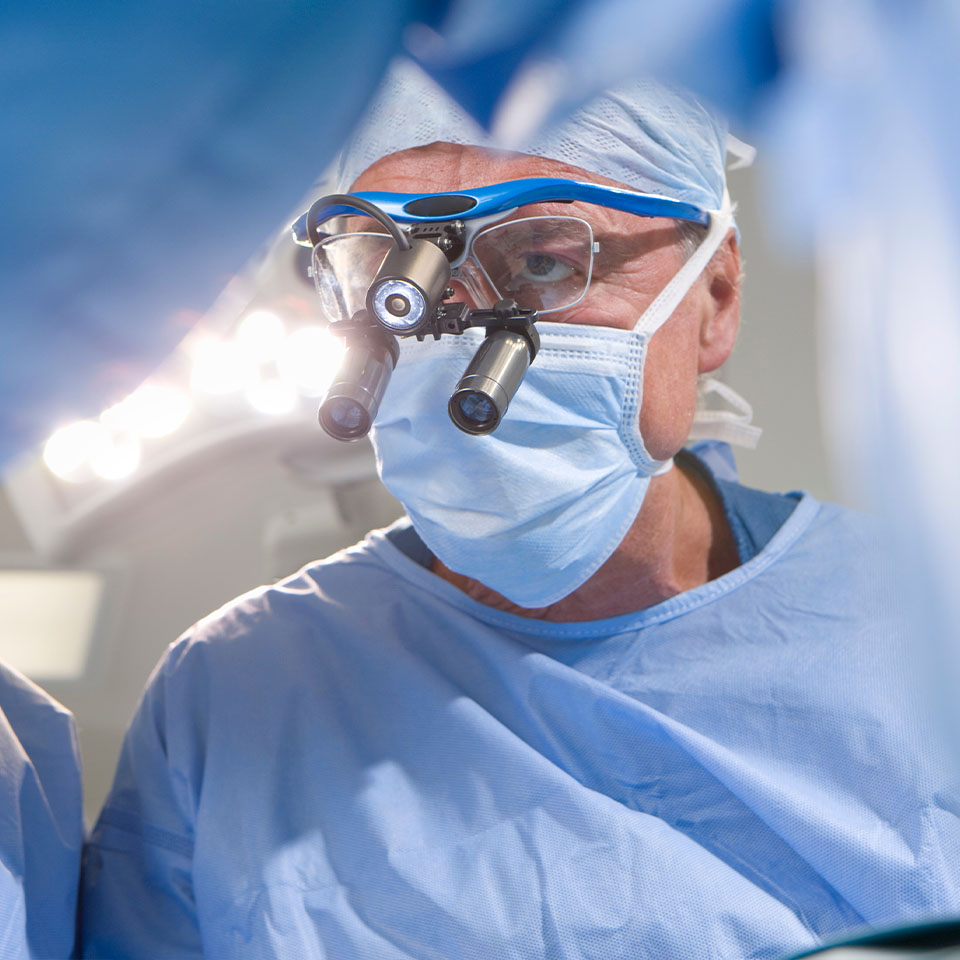 A surgeon wearing surgical scrubs, a face mask, and magnifying loupes performs a procedure under bright operating room lights.