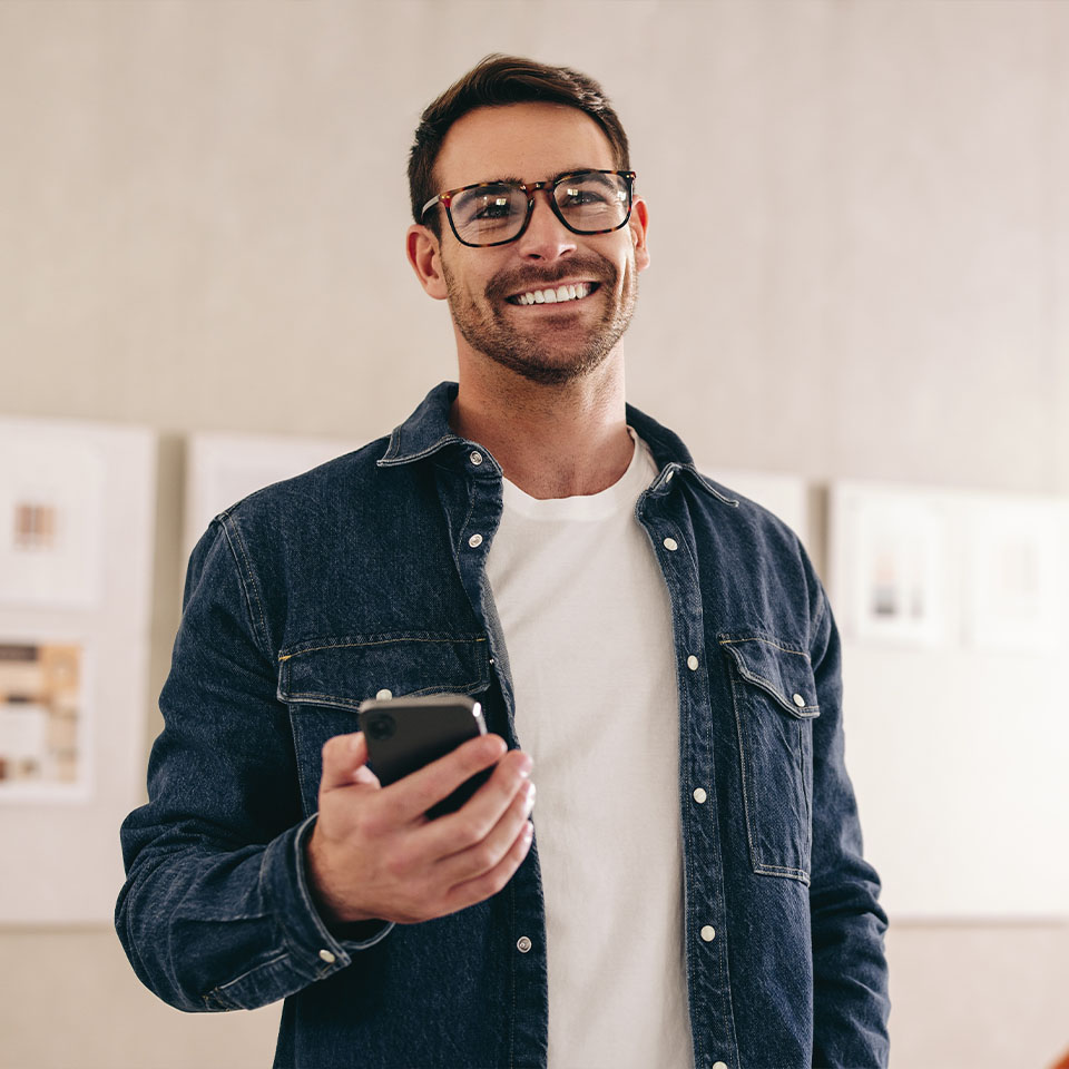 A man wearing glasses and a denim shirt smiles while holding a smartphone, standing indoors with framed pictures on the wall behind him.
