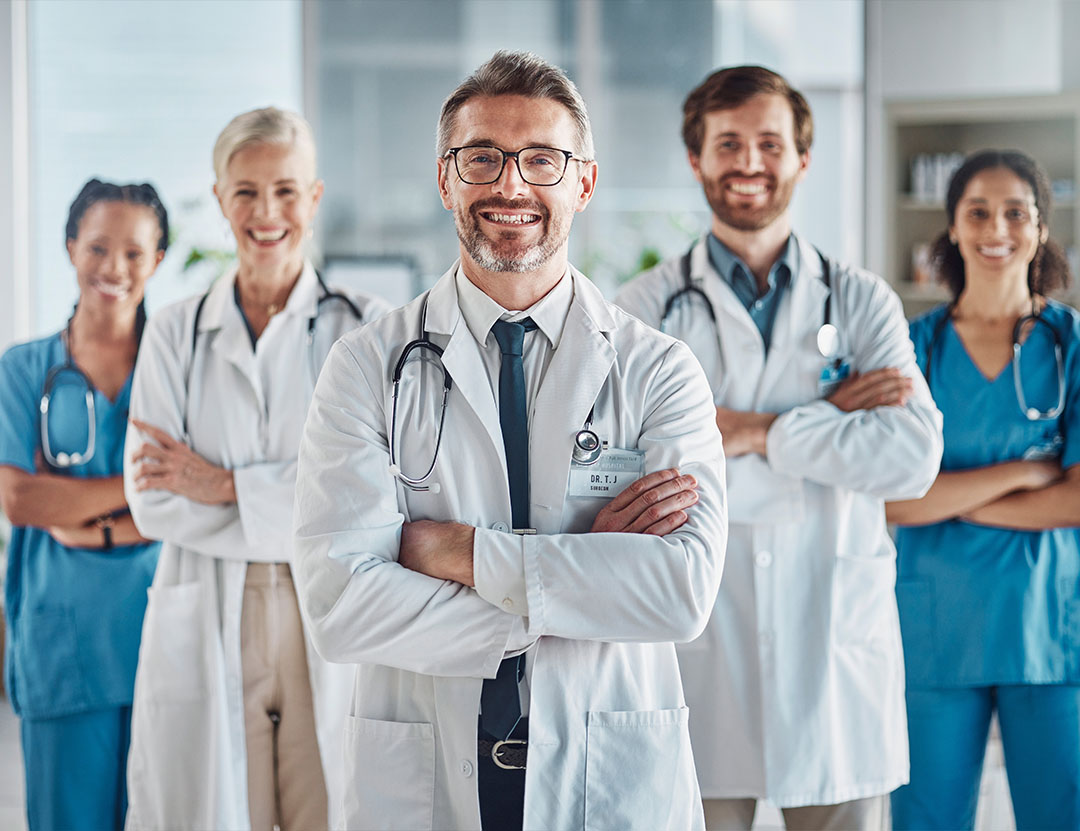 A group of five medical professionals, including doctors and nurses, stand together in a hospital, smiling with arms crossed.