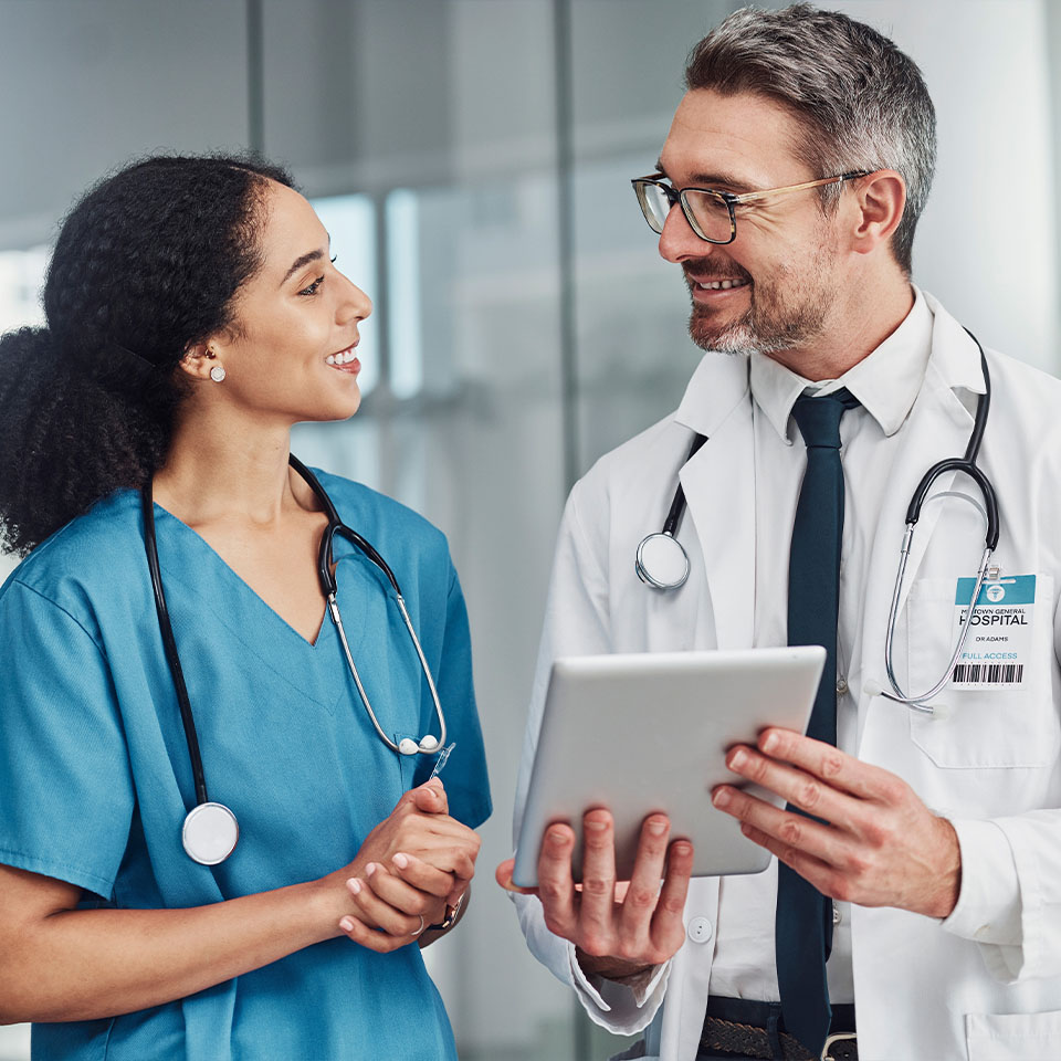 Two healthcare professionals, one in blue scrubs and one in a white coat, stand together indoors, smiling and discussing information on a tablet.