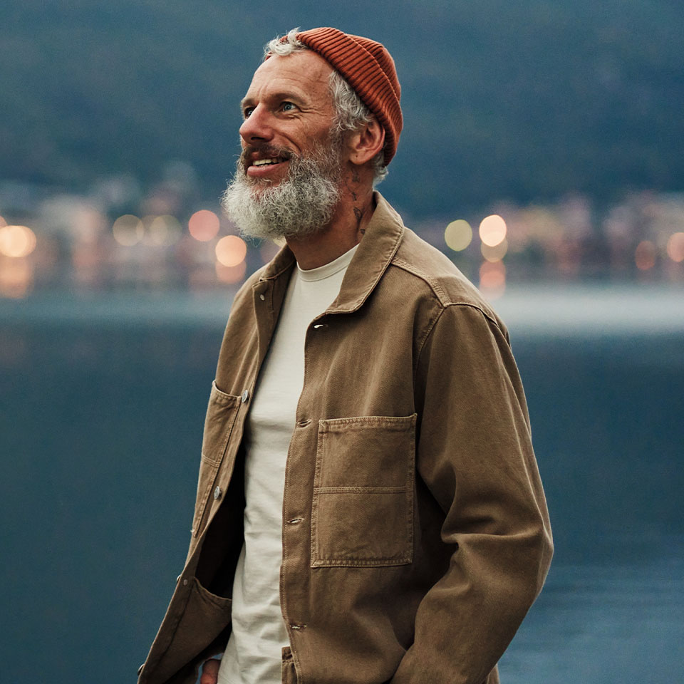 A man with a gray beard wearing a red knit beanie, tan jacket, and white shirt stands outdoors near water with blurred lights in the background.