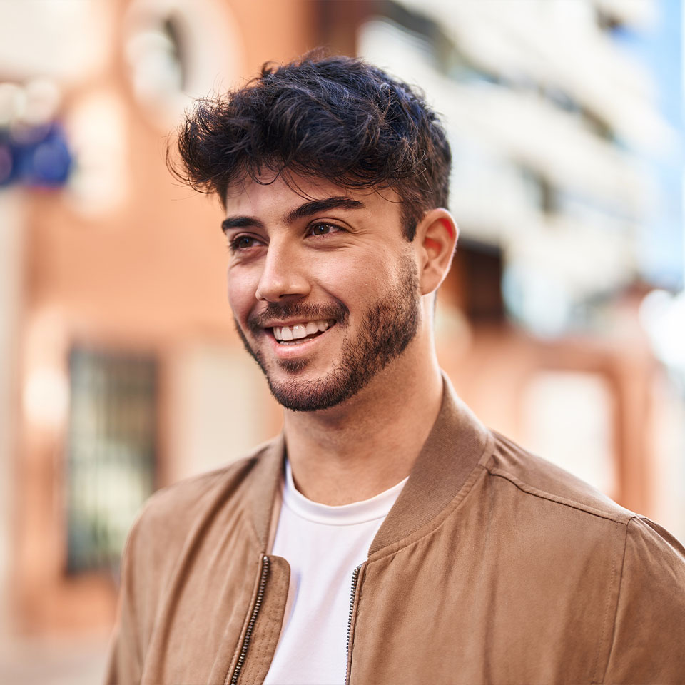 A young man with dark hair and a beard, wearing a light brown jacket and white shirt, smiles outdoors with blurred buildings in the background.