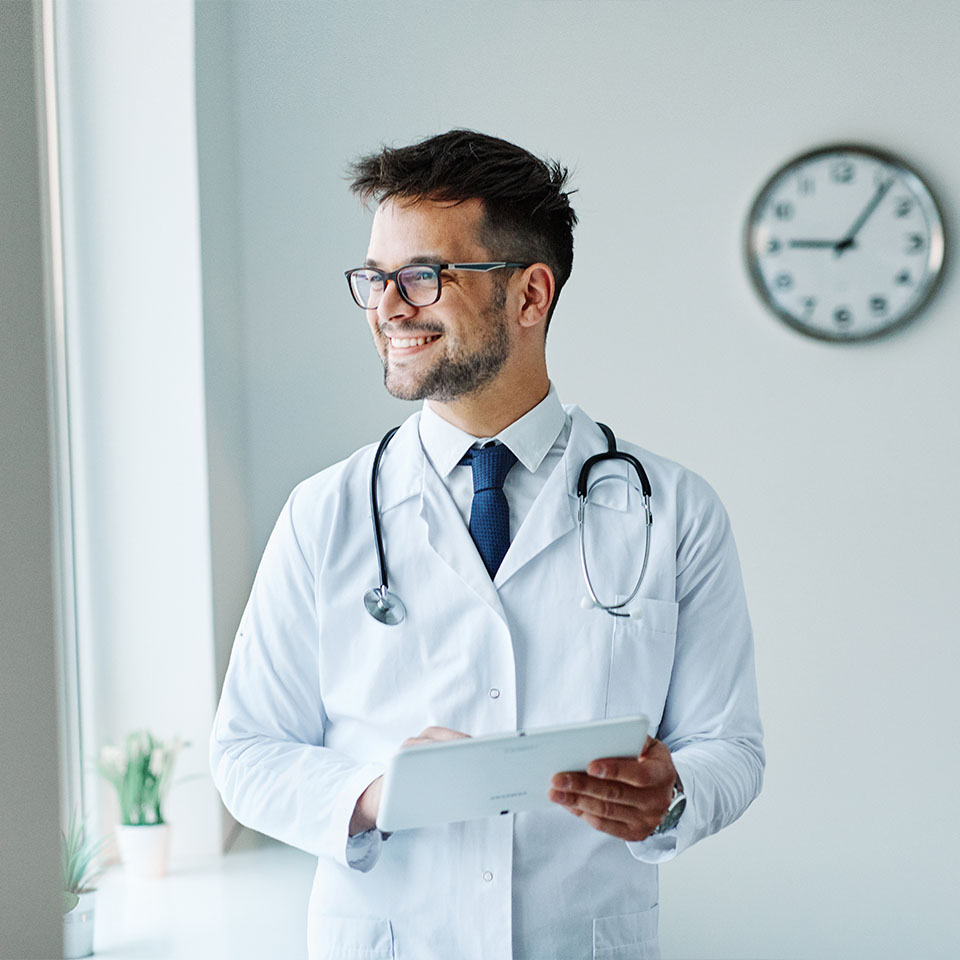 A doctor in a white coat with a stethoscope and tablet stands by a window, smiling. A clock and a small plant are visible in the background.