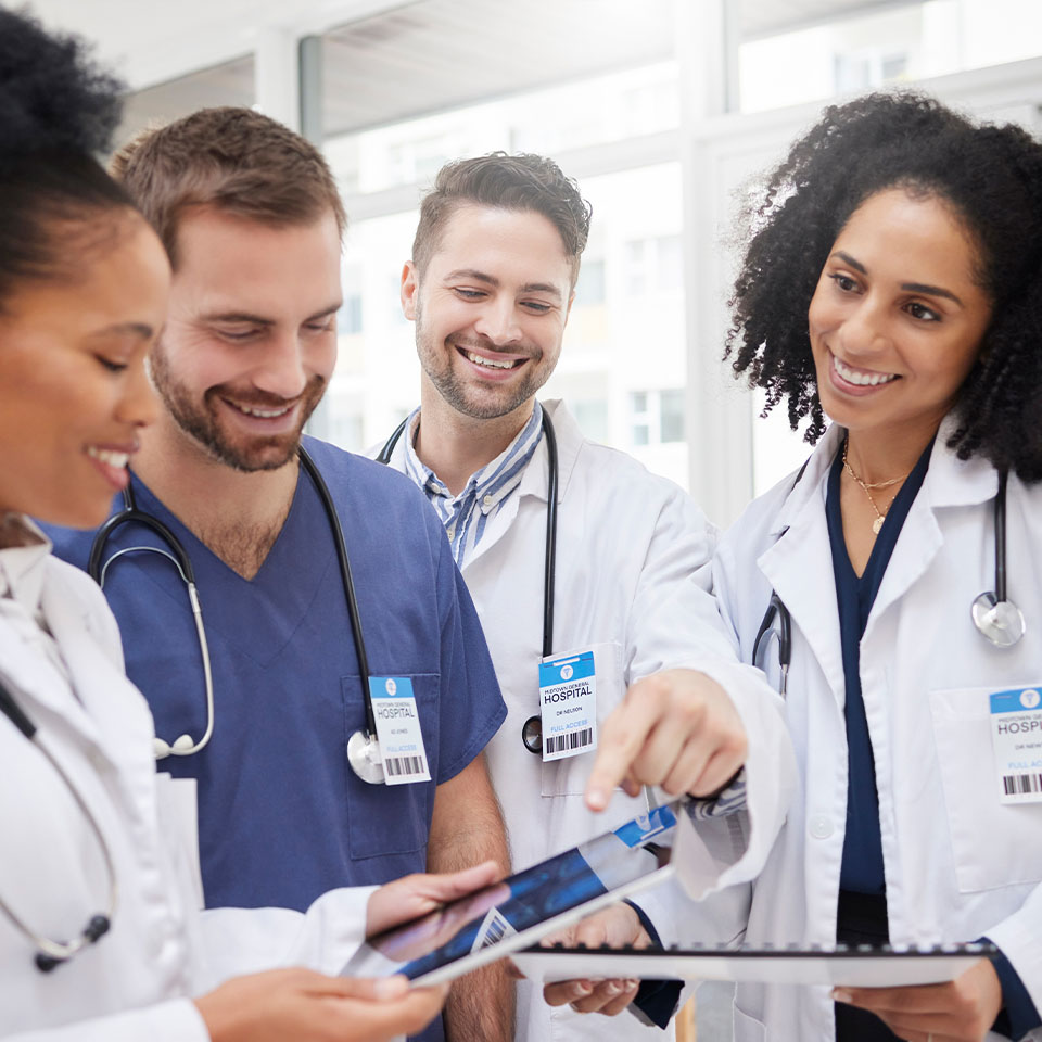 Four healthcare professionals in lab coats and scrubs stand together, smiling and reviewing information on a tablet in a bright hospital setting.