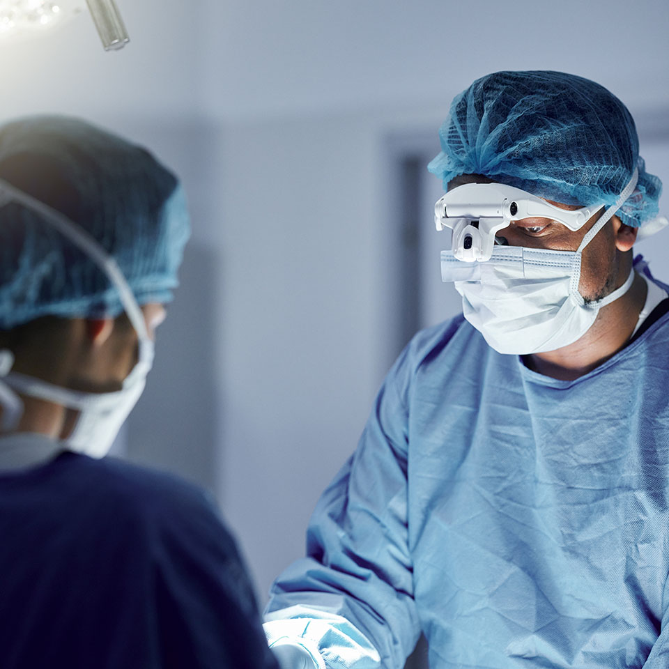Two surgeons in blue scrubs, masks, and hair covers perform a procedure in an operating room under bright surgical lights.