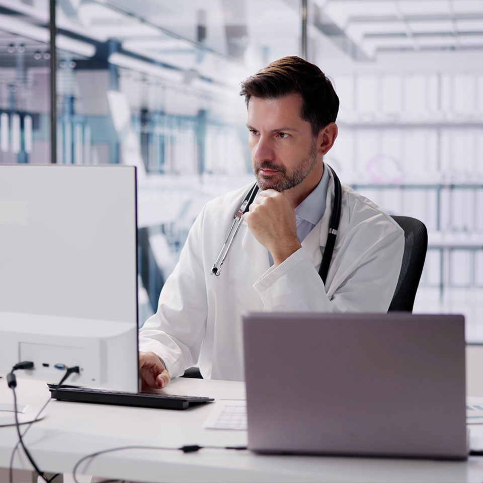 A doctor in a white coat with a stethoscope sits at a desk, looking intently at a computer screen in a modern medical office.