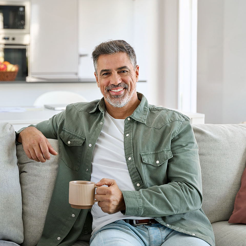 Middle-aged man with gray hair and beard sits on a couch, smiling and holding a beige mug in a modern, bright living room.
