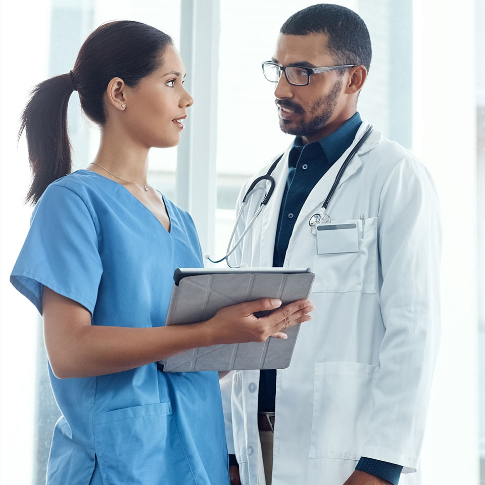 A nurse holding a tablet speaks with a doctor wearing a stethoscope in a hospital setting.