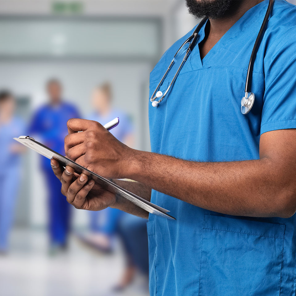 A healthcare professional in blue scrubs writes on a clipboard in a hospital hallway with other staff visible in the background.