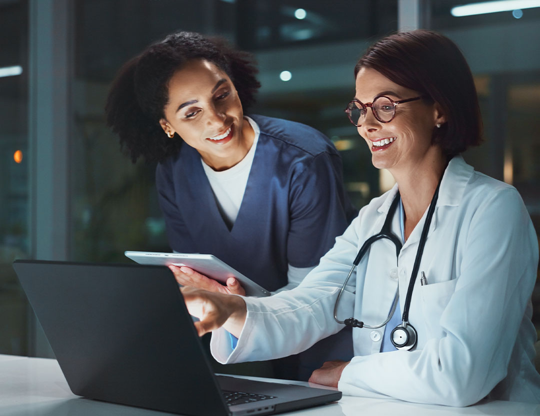 Two healthcare professionals, one in scrubs and one in a lab coat with a stethoscope, review information together on a laptop in a modern medical office.